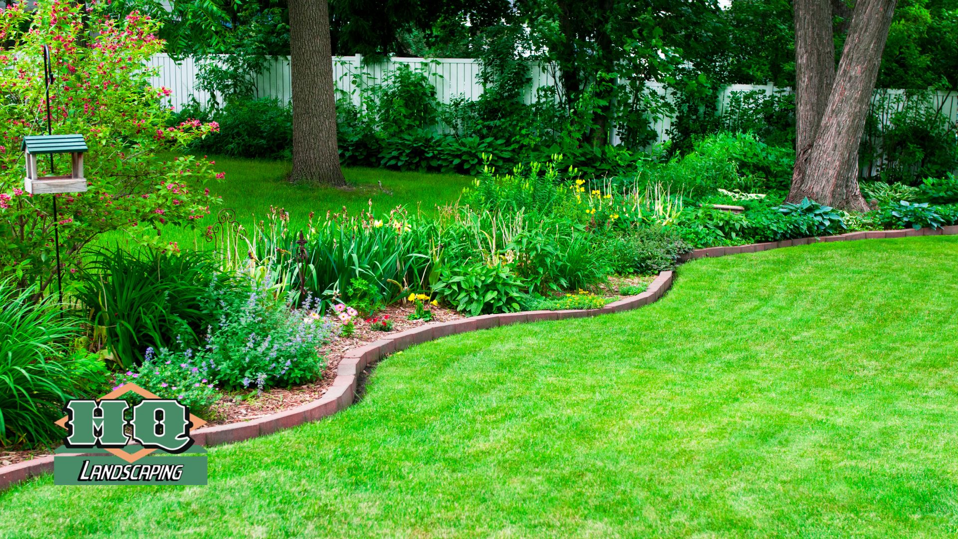 Lawn HQ A beautifully maintained green lawn with a curved flower bed, colorful plants, and a wooden bird feeder, showcasing HQ Landscaping's professional lawn installation and landscaping services.